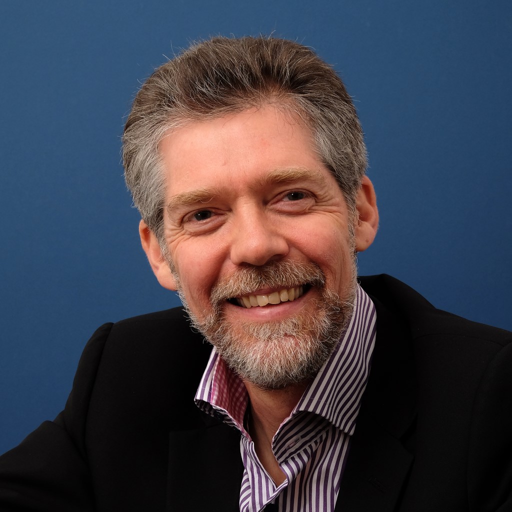 A middle-aged man with short hair and a beard wearing a dark suit and striped shirt