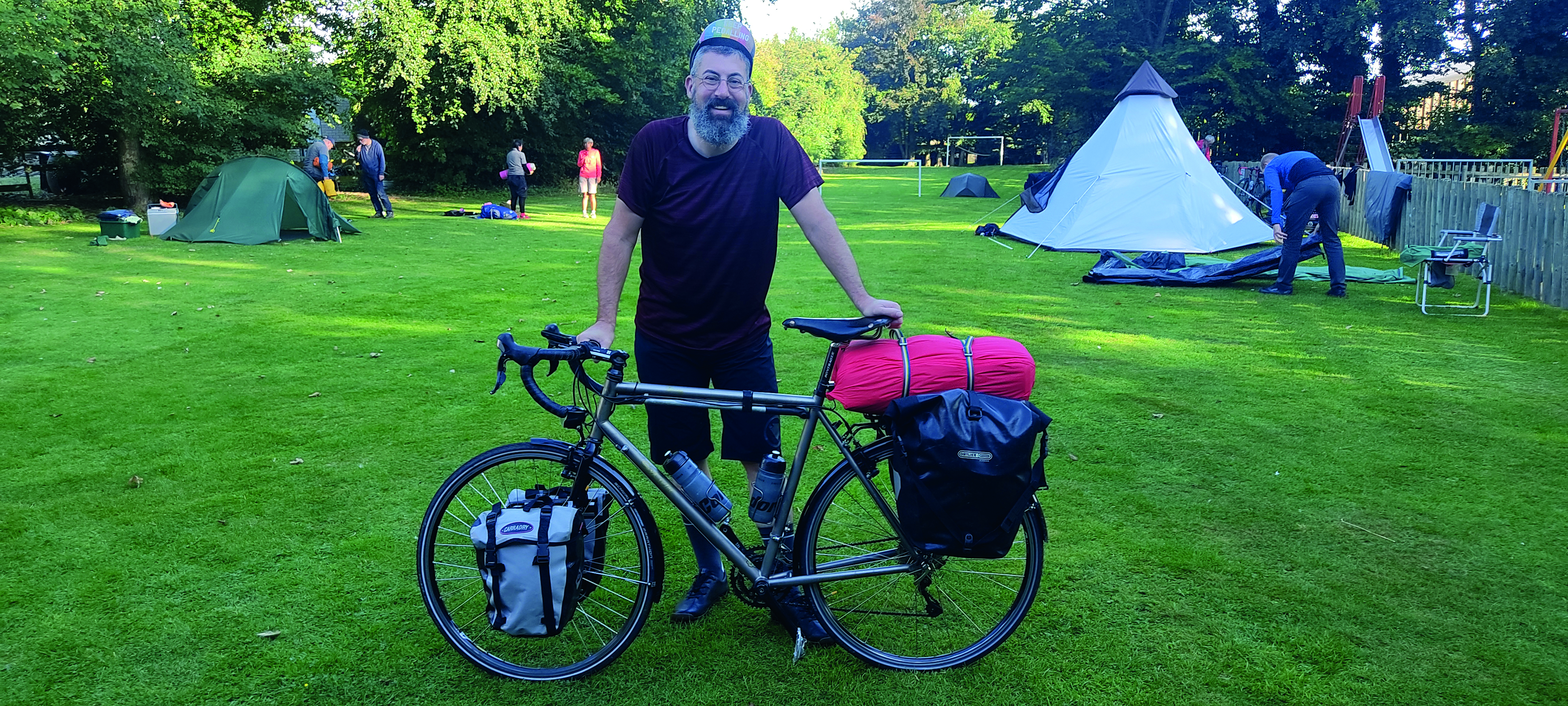 A bearded man stands on a campsite holding his bicycle which is loaded down with panniers