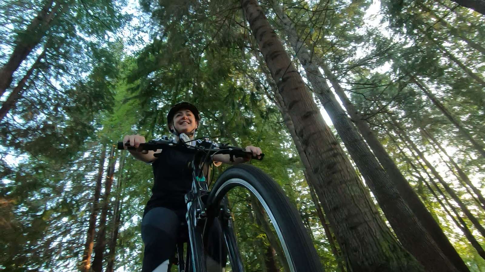 The camera looks up at a woman in dark clothing rides her bicycle through a woodland area