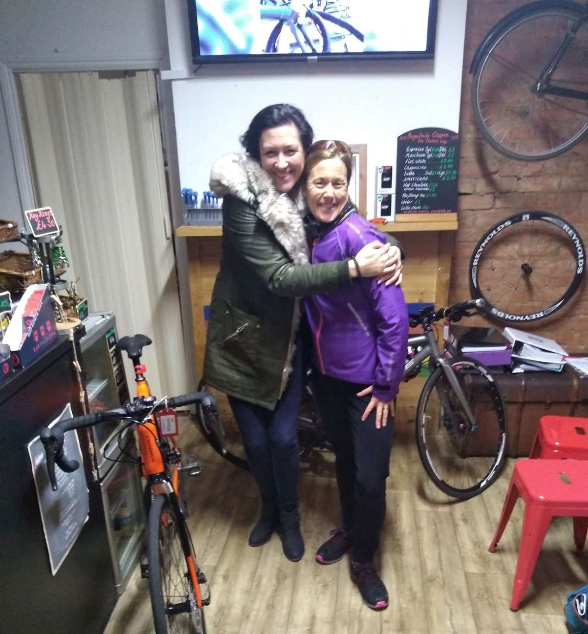 A woman in a green coat and a woman in a purple fleece pictured together in a crowded room with bicycles and wheels lining the walls