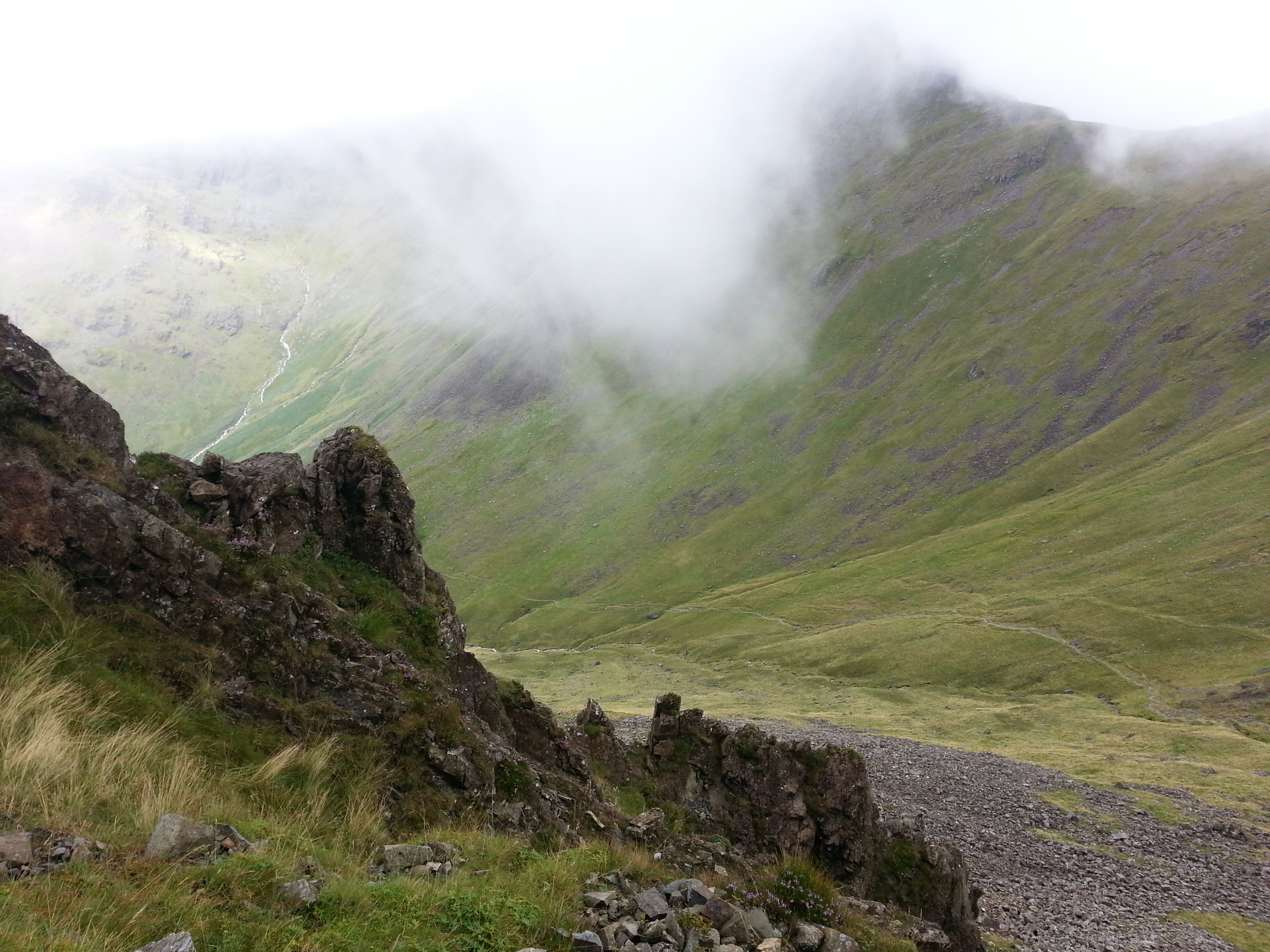 A view of Black Sail Pass, part of Wainwright’s Coast to Coast trail, showing a pass through mountains. A gravel path runs along the bottom of the pass, with grassy mountains rising up on either side. Mist is rolling in.