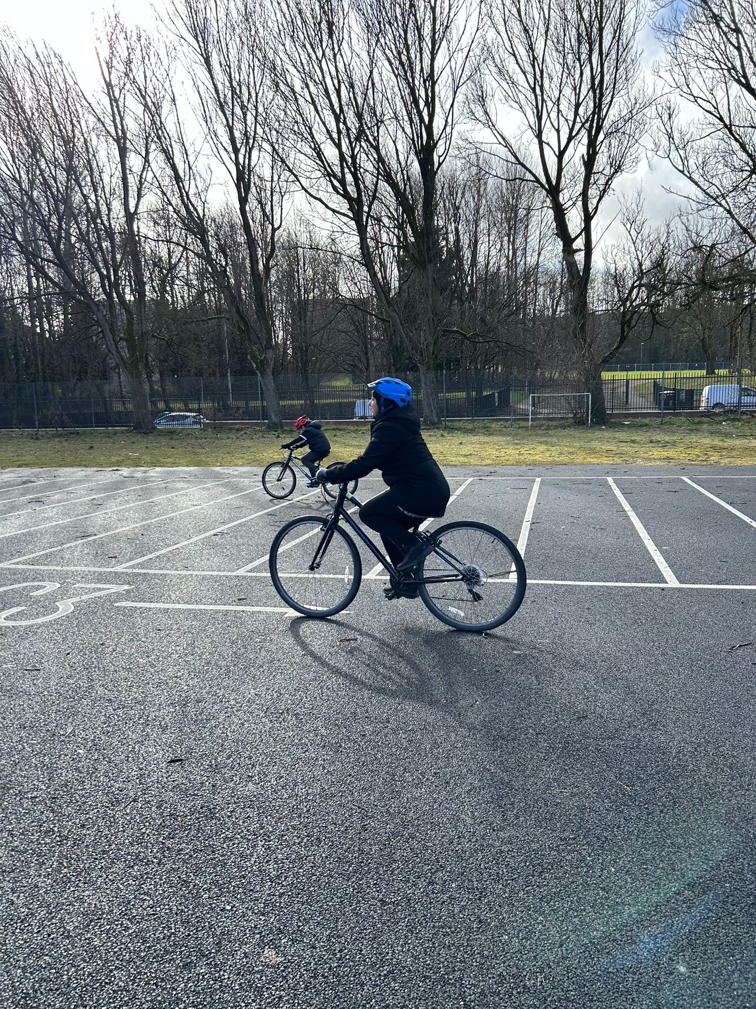 Two cyclists, both wearing helmets and dark clothing, ride through a quiet, wet car park on a grey day, with leafless trees and a football pitch in the background.