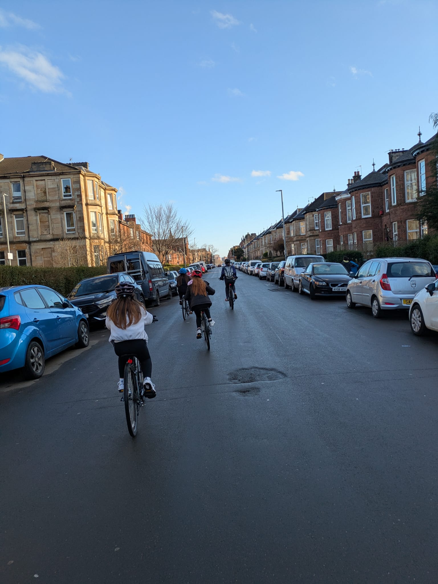 A group of people cycle down a residential street lined with parked cars and traditional tenement buildings on a sunny day.