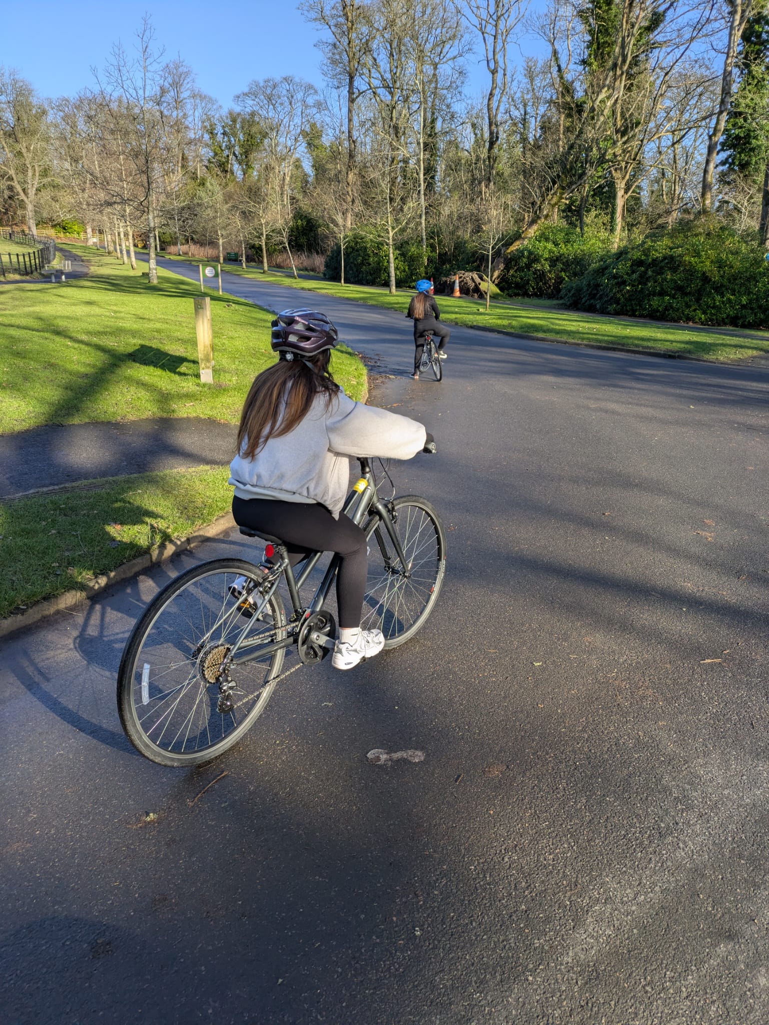 A woman cycles along a quiet, sunlit path in a park, with another cyclist visible in the distance among trees.