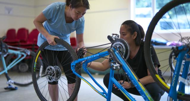 two women standing over a bike indoors. The back wheel is removed and they are working on it