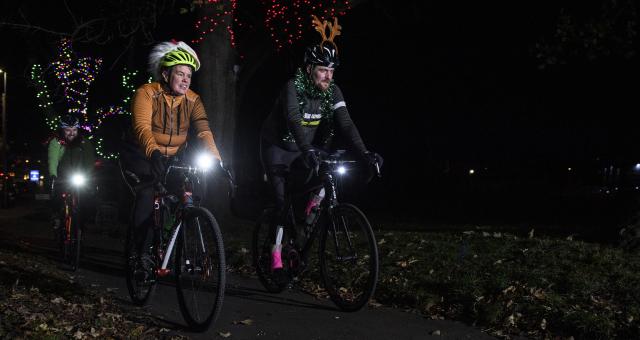 A male and female ride their bikes in the dark with front lights on. One person is wearing a pair of novelty antlers and the other is wearing a Santa hat.
