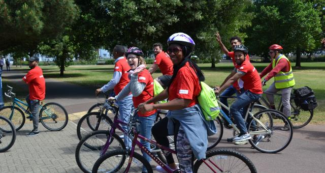 A diverse group of cyclists is cycling around a park. They are wearing matching red T-shirts and are on a variety of cycles.