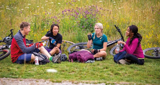 A group of four cyclists are all sitting on the ground having something to eat with their bikes lying next to them