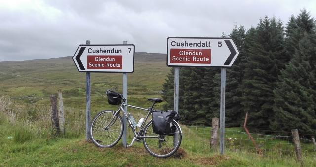 A bike is leaning up against two road signs that are pointing in opposite directions to Cushendun and Cushendall both signs also have the words Glendun Scenic Route in a brown square. In the background is a grassy hill and a line of conifers