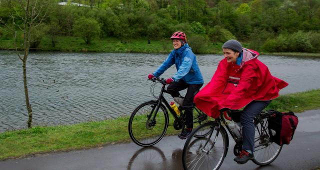 Two women are cycling side by side on a wet path in the rain. One is wearing a blue waterproof, the other a red rain cape. They are both wearing waterproof trousers