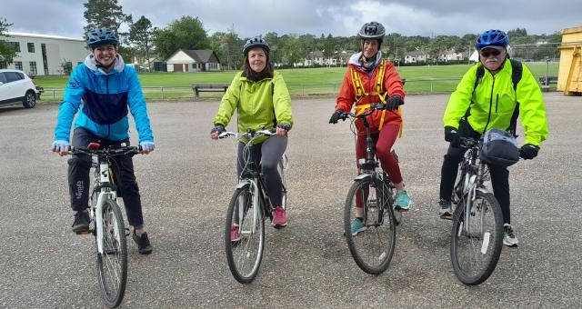 Four people are lined up on their bikes about to set off on a ride. They are wearing brightly coloured jackets and are smiling at the camera.