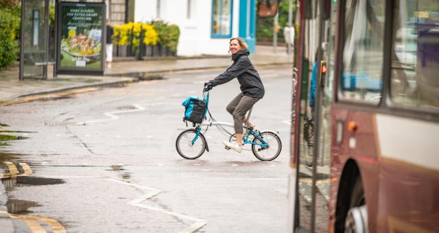 A woman cycles across a road at a pelican crossing in front of a bus. She is riding a Brompton fold-up bike, and is wearing a raincoat and a big smile as she looks at the camera