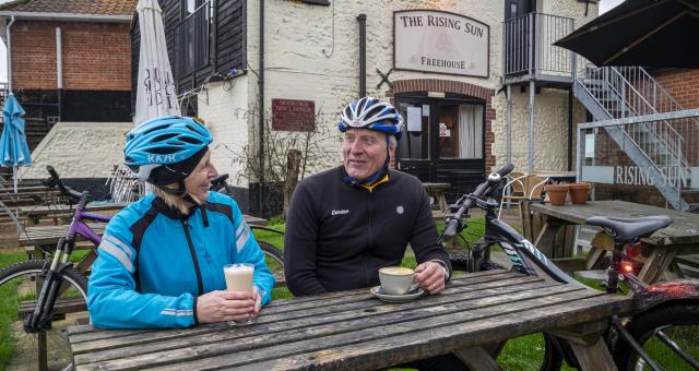 Two cyclists sit at a wooden table in a pub garden, they are holding coffees and chatting
