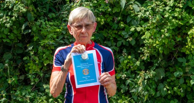 A woman wearing black cycling shorts and a red and blue horizontally striped cycling jersey is holding up a blue certificate.