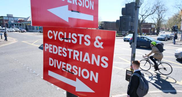 At a set of traffic lights, a red sign with bold white letters reads 'Cyclists and Pedestrians diversion' with an arrow pointing one way. The same sign appears facing the opposite way. In the background a pedestrian waits to cross a busy road