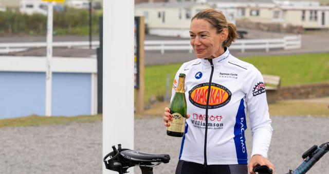 A woman is standing under a John o’ Groats sign. She has a Liv road bike with tri bars and is holding a bottle of champagne, celebrating after completing the Land’s End to John o’ Groats cycle ride.