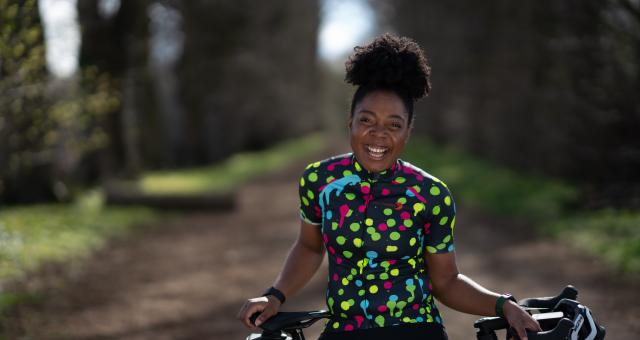 A woman is standing with her road bike. She is perched on the crossbar. She is wearing black cycling shorts and a black and multicoloured polka dot cycling jersey. She is smiling.