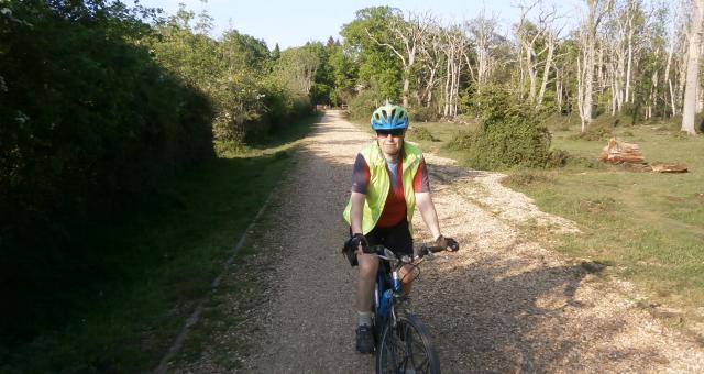 A woman is cycling towards the camera. She is wearing black shorts, an orange and grey jersey and yellow hi-vis jacket, along with a blue and green helmet and sunglasses. The bike is blue. She is cycling on a gravel path bordered by woodland on both sides