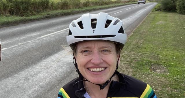 A woman is standing on a grass verge at the side of a road. She is wearing a white cycle helmet and a bright, stripey Stolen Goat cycling jersey. She is smiling at the camera.