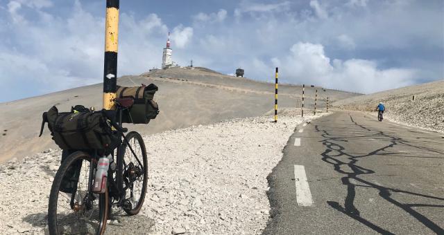 A loaded touring bike is leaning against a pole on a gravel path on a mountain