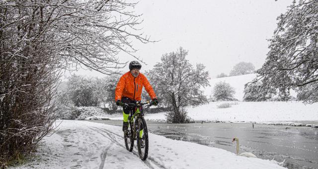 A man is cycling along a snowy path next to a frozen river. It’s a very wintry scene, it’s snowing. He is wearing a bright orange winter jacket and black winter cycling leggings with bright yellow sections. There’s a single swan on the river