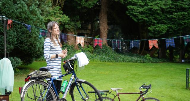 A woman is standing on a makeshift stage. She has a Dawes touring bike with her; an open notebook is balanced on the bike’s handlebars. She is wearing a blue and white striped top, black three-quarter-length trousers and cycling shoes. She is smiling.