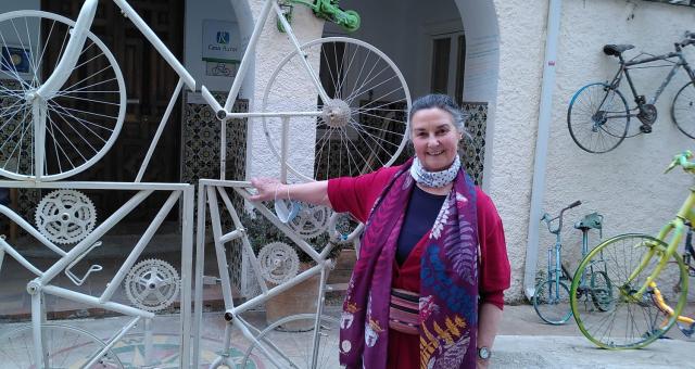 A woman is standing at the gates of a hotel in Spain. The building is covered with old bicycles and even the gates are made from bike frames. She is wearing black trousers, a bright cerise cardigan and blue top, as well as a brightly patterned scarf