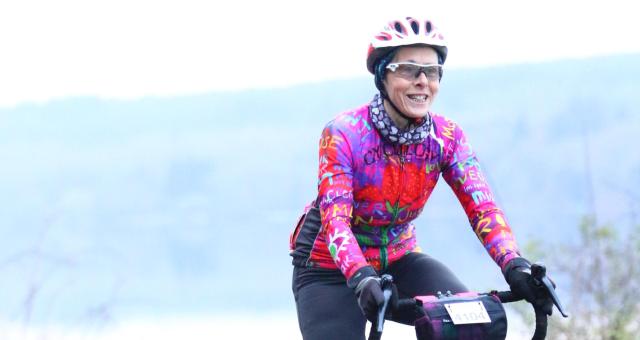 A woman is cycling along the road. She is on a road bike which has a number attached to a handlebar bag; she is taking part in an organised cycling event. She is wearing black cycling leggings and a very bright multicoloured jersey