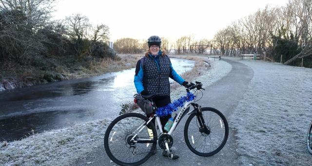 A woman is standing with a light grey Trek mountain bike. It’s winter. She is wearing a warm winter jacket and trousers. Her bike is decorated with blue tinsel. There is frost on the pavement, grass and trees and the stream behind her has ice on it