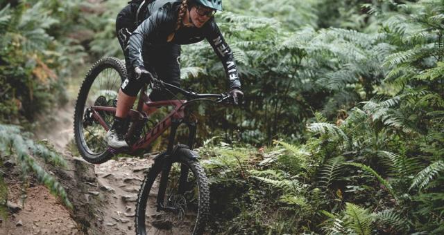 A woman is cycling downhill through a forest. She is riding a Scott mountain bike. She is wearing black three-quarter-length cycling leggings and a black jacket, as well as a green helmet. Credit: Roo Fowler