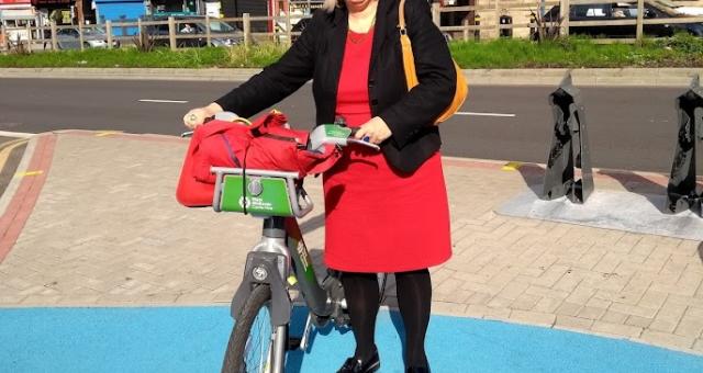 A woman is standing with a hire bike on some of Birmingham’s cycle infrastructure, the blue routes. She is wearing a red dress, black cardigan and black tights. She has a yellow handbag over her shoulder and a red bag in the bike’s basket