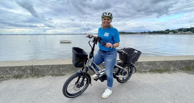 Lucie on a silver e-cycle with front and back baskets, standing in front of Poole harbour. She is wearing blue jeans and a blue BH Active Travel T-shirt