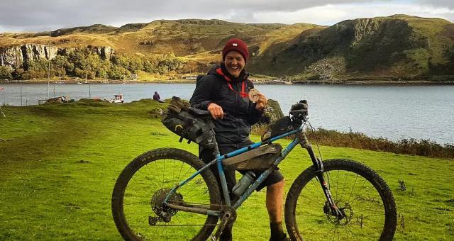 A woman is standing with a very muddy bike and holding a medal, having just completed an off-road triathlon on the Isle of Kerrera. She is smiling broadly