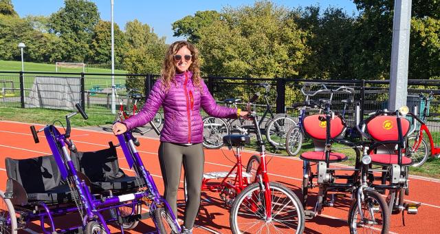 A woman is standing on a racetrack. She is wearing black leggings and a violet padded jacket, along with sunglasses. She has long, wavy, dark blonde hair. She is surrounded by various non-standard cycles, showing the different options for different needs