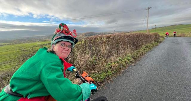A woman is riding a bike while wearing a Christmas elf outfit. She is looking back at the camera and pulling a funny face