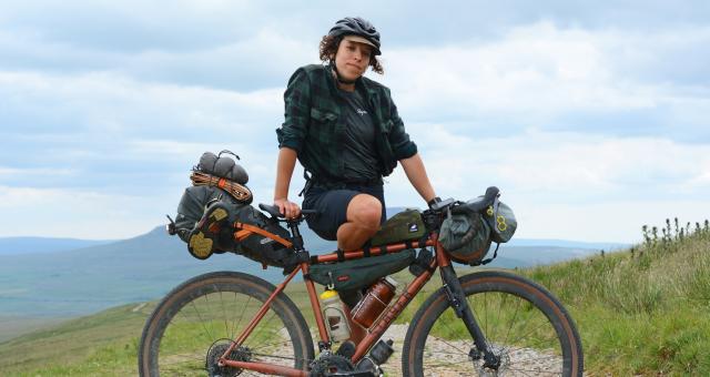 A woman is posing with a heavily laden bikepacking bike. She is wearing black shorts, a black Rapha T-shirt and check shirt, and a black cycling helmet with a cap underneath. She is in a remote location with mountains in the background