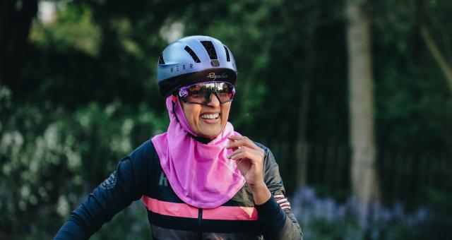 A woman in cycling gear is smiling at the camera. She is wearing a blue cycling helmet with a Rapha cap underneath. She has on cycling sunglasses, a stripey Rapha cycling jersey and a bright pink headscarf
