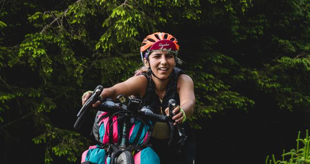 A woman is pushing her bike uphill. Only the bike’s handlebars and top of the front wheel are in shot, along with packed bikepacking bags. She is wearing an orange helmet and a Rapha cap