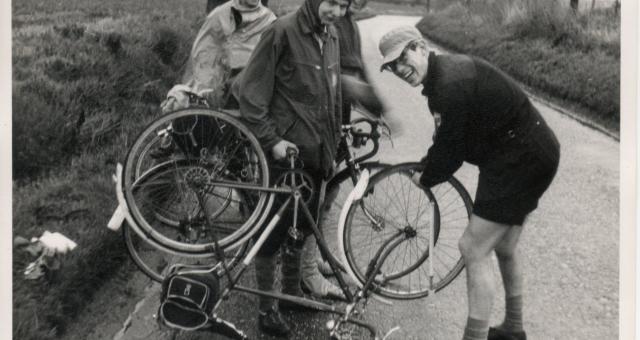 A scan of an old black and white photo showing a group of men with bikes. One bike is turned over and propped up on its saddle as one of the men takes off the front wheel