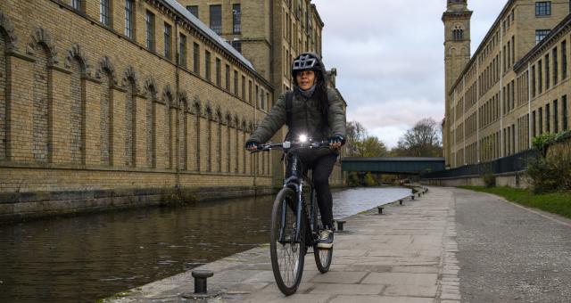 A woman is cycling along a canal tow path in the evening. She has a bright front light on her bike.