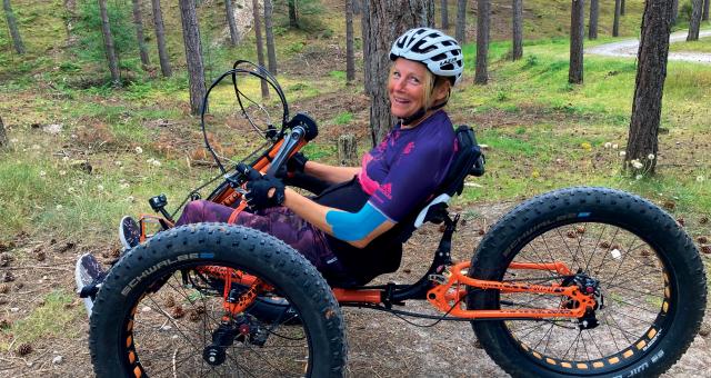 A woman is riding an orange fat trike handcycle through woodland. She is wearing purple kit and a white helmet. She has long blonde hair in a ponytail and is smiling at the camera.
