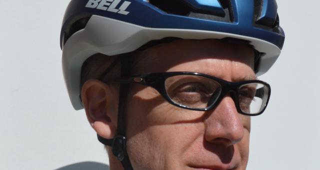 A man in glasses is wearing a blue and grey cycling helmet with the Bell logo on the side