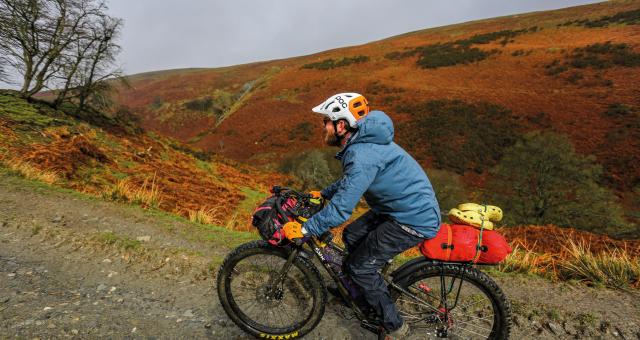 A man is riding up a slope on a gravel path through a very hilly landscape. He's wearing waterproofs and there is rain on his jacket