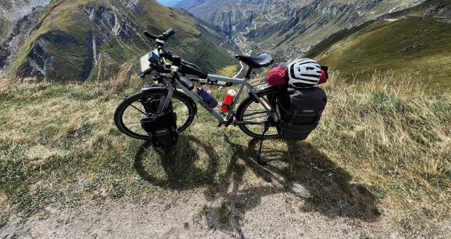 A loaded flatbar touring bike with front and rear panniers, saddlebag and top tube bag is propped up at the top of a mountain with a valley laid out behind it