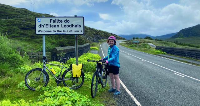 A woman is standing with her bike on the side of a road next to a road sign. She is wearing cycling shorts and helmet and a waterproof. Another bike with a packed pannier is leaning against the sign. The sign reads Failte do dh'Eilean Leodhais, with the English translation underneath: Welcome to the Isle of Lewis