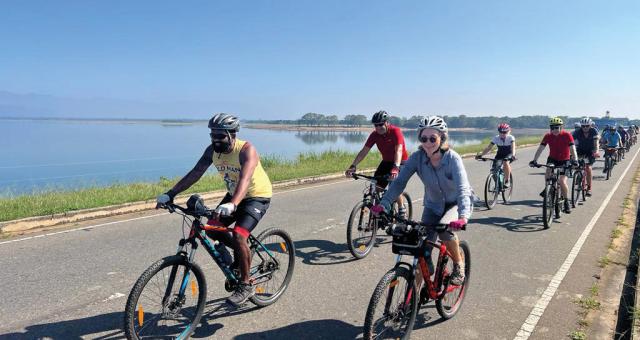 A group of people is cycling along a tarmac road in Sri Lanka. They are in a mix of cycling and non-cycling clothing, all are wearing helmets. There's a huge lake in the background.