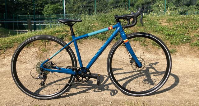 A blue gravel bike propped up on a gravel track. It has pale blue, red, yellow and orange rings painted around the frame