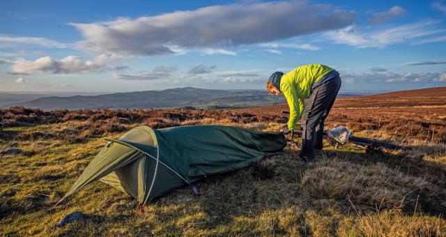 A man is setting up a bivvy bag in the countryside. He is wearing waterproof trousers and bright yellow waterproof jacket. A bike is lying on the ground in the background