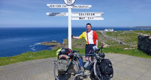 A man is standing next to the Land's End sign, which points to the Scilly Isles and John o' Groats. It also says Saltdean and the date May 21st. He is wearing shorts and a club cycling jersey. He is leaning on the sign. He has a loaded touring bike.