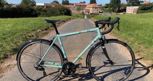 A pale turquoise road bike with mudguards propped up on a narrow country lane